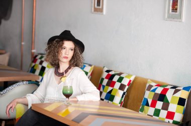 Siyah şapkalı mutlu esmer kız barın içinde oturmuş Uzağa doğru gülümserken bir bardak soğuk yeşil meyve suyu içiyor. Bankta meyve suyu içen beyaz gömlekli güzel bir kadın. 