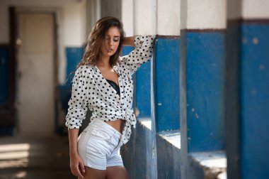 Beautiful girl with white shirt and white shorts posing in old hall with columns blue painted. Attractive long hair brunette side view against ancient pillars. Dark hair young woman with gorgeous eyes