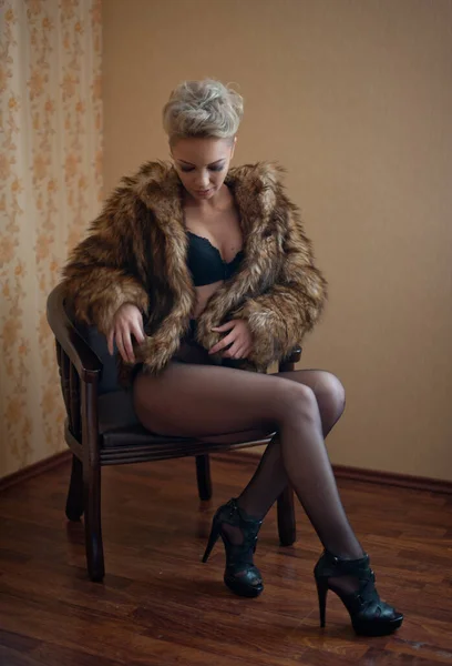 椅子に挑発的にポーズをとるパンスト 黒ブラと毛皮のコートと魅力的なブロンドモデル 官能的なブロンドのファッションの肖像画 スタジオショット 官能的な女性で黒ランジェリーポーズ反対壁 — ストック写真