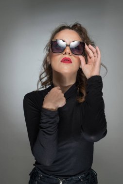 Gri arka planda siyah bluzlu ve güneş gözlüklü bir kadın. Kırmızı dudaklı güzel bir kız. Güneş gözlüklü çekici bir kadının portresi. Kameraya bak.
