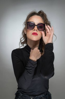 Gri arka planda siyah bluzlu ve güneş gözlüklü bir kadın. Kırmızı dudaklı güzel bir kız. Güneş gözlüklü çekici bir kadının portresi. Kameraya bak.