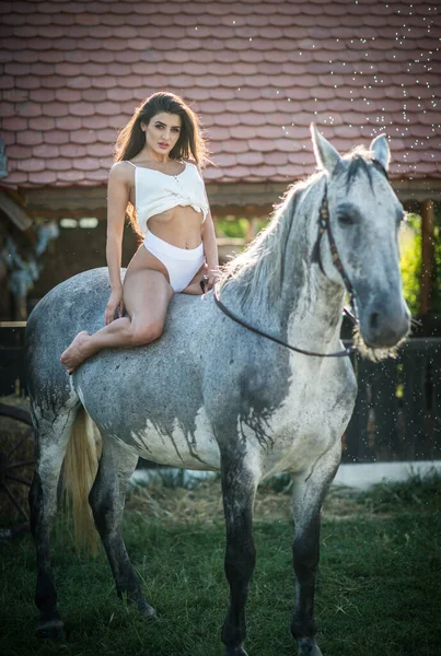 長髪の美しいブルネット女性 白いビキニと白いシャツは 水滴の下で白い馬に乗っています ランチで馬と交流して楽しむ 官能的で魅力的な女の子 — ストック写真