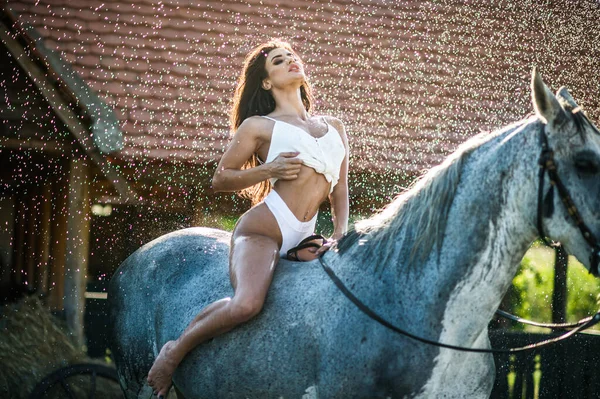美丽的黑发女人 白色比基尼和白色衬衫骑在水滴下的白马上 一个性感迷人的女孩和一匹马在农场里嬉戏玩耍 — 图库照片