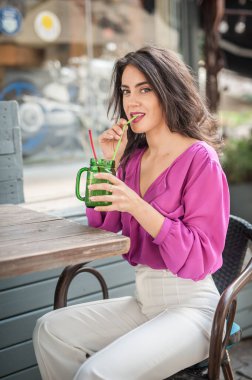 Barda oturmuş, kameraya gülümserken bir bardak limonata içen pembe bluzlu mutlu esmer kız. Sandalyede beyaz pantolon giymiş, meyve suyu içen genç, seksi bir kadın.