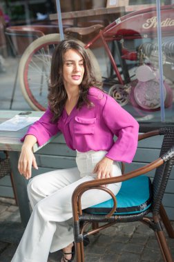 Barda oturmuş, kameraya gülümserken bir bardak limonata içen pembe bluzlu mutlu esmer kız. Sandalyede beyaz pantolon giymiş, meyve suyu içen genç, seksi bir kadın.