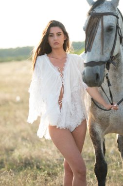 Uzun saçlı güzel esmer kadın, beyaz şeffaf bluz, beyaz bikini ve at. Bir kızın portresi. Şehvetli ve çekici bir kız bir atla etkileşime geçip eğleniyor.