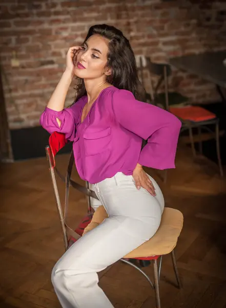 漂亮的年轻的黑发女人 穿着紫色的衬衫和白色的裤子 靠着砖墙在室内摆姿势 一个年轻貌美的黑发女子的画像 图库图片