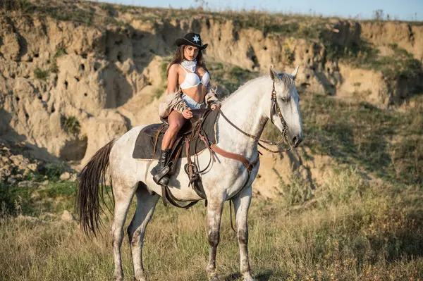 一个性感的女牛仔骑着马在沙漠里戴着牛仔帽骑着马的漂亮姑娘 美丽的女牛仔骑着马在山上 户外生活方式肖像 图库图片
