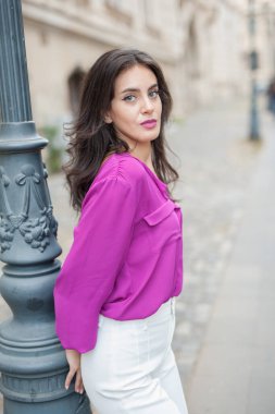 Pembe ceketli, beyaz pantolonlu, sokakta poz veren güzel esmer bir kadın. Mor bluzlu ve beyaz pantolonlu güzel bir kadının portresi. Sokakta bir kafede poz veriyor.