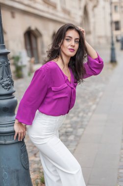 Pembe ceketli, beyaz pantolonlu, sokakta poz veren güzel esmer bir kadın. Mor bluzlu ve beyaz pantolonlu güzel bir kadının portresi. Sokakta bir kafede poz veriyor.