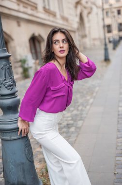 Pembe ceketli, beyaz pantolonlu, sokakta poz veren güzel esmer bir kadın. Mor bluzlu ve beyaz pantolonlu güzel bir kadının portresi. Sokakta bir kafede poz veriyor.