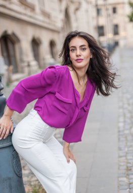 Pembe ceketli, beyaz pantolonlu, sokakta poz veren güzel esmer bir kadın. Mor bluzlu ve beyaz pantolonlu güzel bir kadının portresi. Sokakta bir kafede poz veriyor.