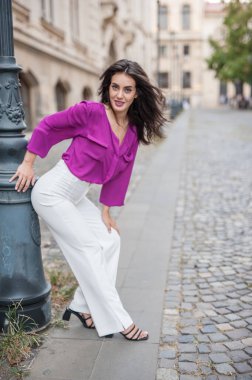 Pembe ceketli, beyaz pantolonlu, sokakta poz veren güzel esmer bir kadın. Mor bluzlu ve beyaz pantolonlu güzel bir kadının portresi. Sokakta bir kafede poz veriyor.