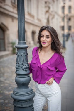 Mor bluzlu ve beyaz pantolonlu güzel bir esmer kadın bir sütuna yaslanıyor. Mor ceketli güzel bir esmer kadının portresi. 