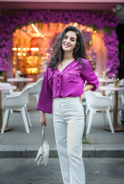 Young beautiful brunette woman in a pink jacket and white pants posing on the street. Portrait of a beautiful young woman in a purple blouse and white pants posing in an outdoor cafe on the street