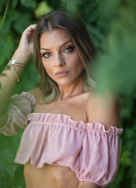 Göz alıcı yeşil gözlü güzel bir kadının portresi, açık hava ortamında çekilmiş. Çerçevesi yemyeşil yeşilliklerle süslenmiş. Açık pembe omuzlu ve etekli bir kadın poz veriyor.