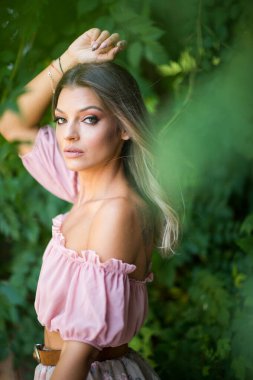 Göz alıcı yeşil gözlü güzel bir kadının portresi, açık hava ortamında çekilmiş. Çerçevesi yemyeşil yeşilliklerle süslenmiş. Açık pembe omuzlu ve etekli bir kadın poz veriyor.