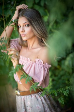 Göz alıcı yeşil gözlü güzel bir kadının portresi, açık hava ortamında çekilmiş. Çerçevesi yemyeşil yeşilliklerle süslenmiş. Açık pembe omuzlu ve etekli bir kadın poz veriyor.