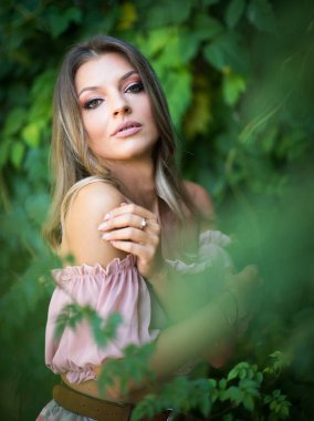 Göz alıcı yeşil gözlü güzel bir kadının portresi, açık hava ortamında çekilmiş. Çerçevesi yemyeşil yeşilliklerle süslenmiş. Açık pembe omuzlu ve etekli bir kadın poz veriyor.