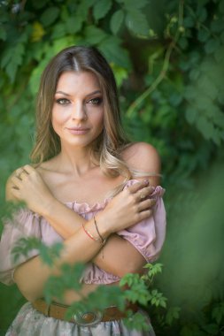Göz alıcı yeşil gözlü güzel bir kadının portresi, açık hava ortamında çekilmiş. Çerçevesi yemyeşil yeşilliklerle süslenmiş. Açık pembe omuzlu ve etekli bir kadın poz veriyor.