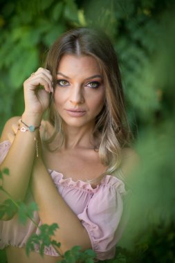 Göz alıcı yeşil gözlü güzel bir kadının portresi, açık hava ortamında çekilmiş. Çerçevesi yemyeşil yeşilliklerle süslenmiş. Açık pembe omuzlu ve etekli bir kadın poz veriyor.