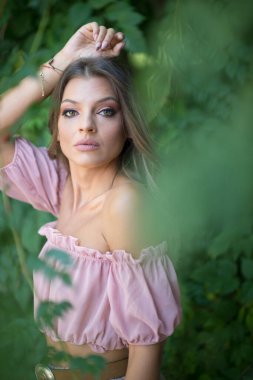 Göz alıcı yeşil gözlü güzel bir kadının portresi, açık hava ortamında çekilmiş. Çerçevesi yemyeşil yeşilliklerle süslenmiş. Açık pembe omuzlu ve etekli bir kadın poz veriyor.