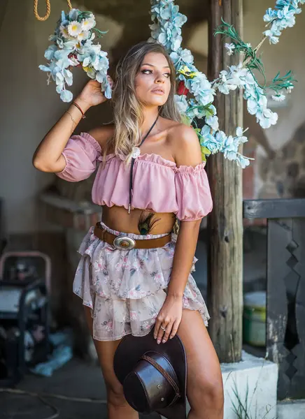 Pembe bir boho hasat bluzu ve çiçekli etek giymiş şık bir genç kadın kırsal alanda el yapımı bir çiçek salıncağının yanında güven içinde poz veriyor. Sahne yumuşak doğal ışıkla yıkanır.