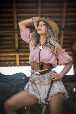 Şık bir batı portresi, omuzları açık pembe bir kadın ve çiçekli bir etek, kırsal bir ahşap önünde bir tüfek tutuyor kahverengi çizmeleri ve kırsal bir arka planı var.