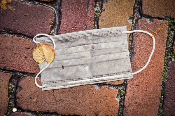 Wet facemask and fallen leaf on the bricks of a pavement in autumn