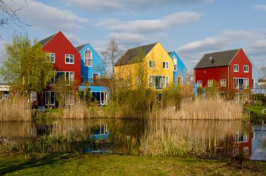 Culemborg, The Netherlands, April 8, 2023: Scandinavian style houses with wooden facades in primary colors reflect in a pond in the golden hour on an evening in spring clipart