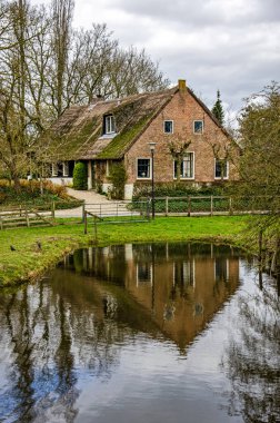 Noordeloos, 13 Mart 2025: Sazdan çatılı eski çiftlik evi ve nehre yansıyan tuğla cephe