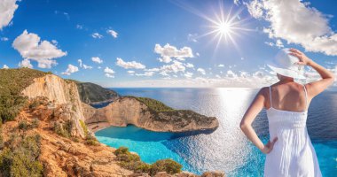Beyaz yaz elbisesi içinde güzel bir kadın bir uçurumun kenarında duruyor ve Yunanistan 'ın Zakynthos adasındaki ünlü gemi enkazı plajı Navagio' nun manzarasının tadını çıkarıyor.