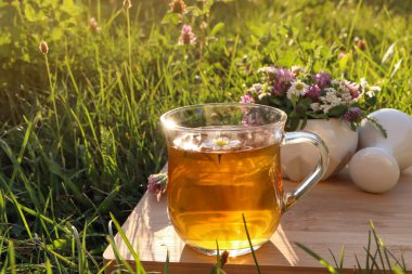 Bir fincan aromatik bitkisel çay, tokmak ve seramik havan, yeşil çimlerin üzerinde farklı kır çiçekleri.