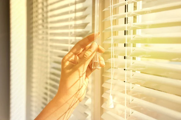 Woman opening horizontal blinds on window indoors, closeup