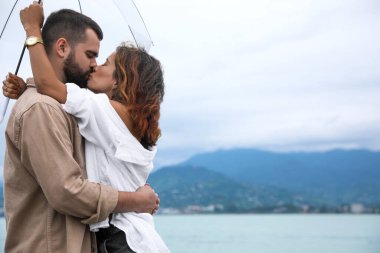 Şemsiyeli genç çift kumsalda yağmurun altında eğleniyor, mesajlaşmak için yer var.