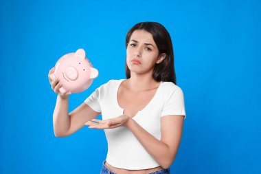 Açık mavi arka planda domuz kumbarası olan üzgün genç bir kadın.