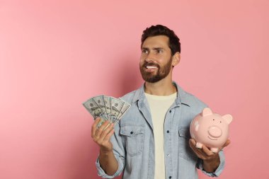 Parası ve kumbarası olan mutlu bir adam soluk pembe arka planda.