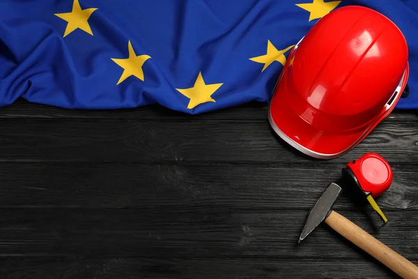 Red hard hat, measuring tape, hammer and flag of European Union on wooden black table, flat lay. Space for text