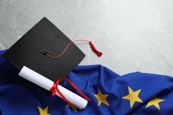 stock image Black graduation cap, diploma and flag of European Union on light grey marble table, flat lay. Space for text