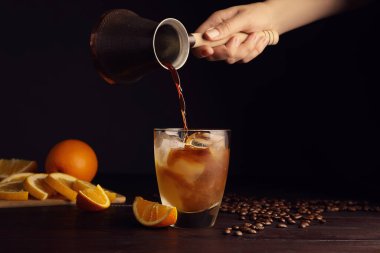 Woman pouring coffee into glass with orange juice and ice cubes at wooden table, closeup