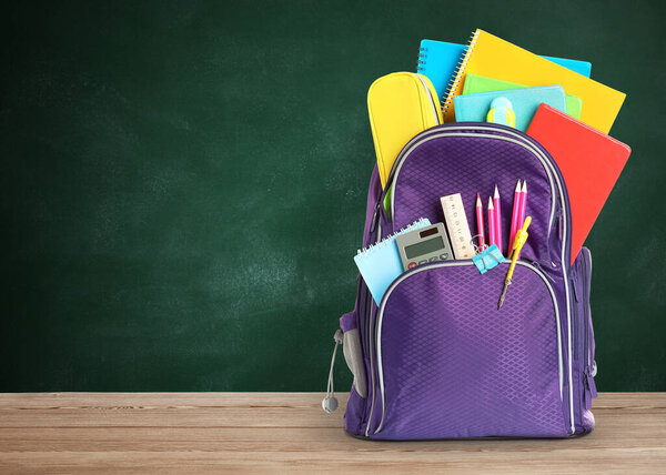 Backpack with school stationery on wooden table near green chalkboard, space for text