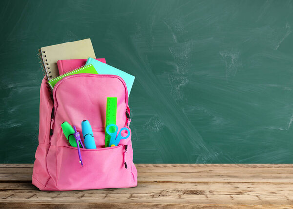 Backpack with school stationery on wooden table near green chalkboard, space for text