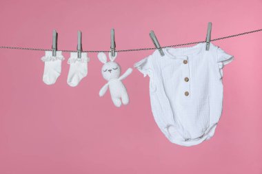Farklı bebek kıyafetleri ve pembe arka planda çamaşır ipinde kuruyan tavşan oyuncağı.