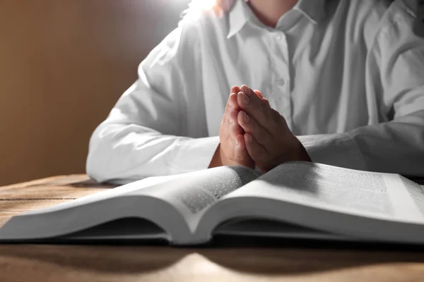 Woman holding hands clasped while praying over Bible at wooden table, closeup