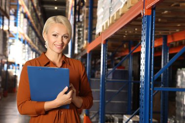 Happy manager holding clipboard in warehouse with lots of products clipart