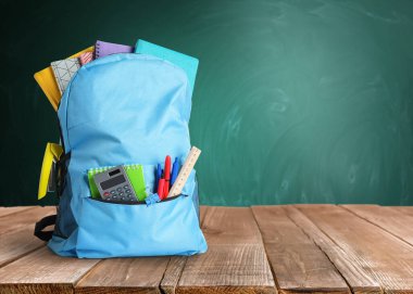 Yeşil karatahtanın yanındaki ahşap masanın üzerinde okul kırtasiyesi olan sırt çantası, metin için alan