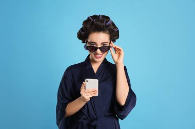 Açık mavi arka planda akıllı telefon kullanan ipek bornozlu, saç bigudili ve güneş gözlüklü mutlu bir genç kadın.