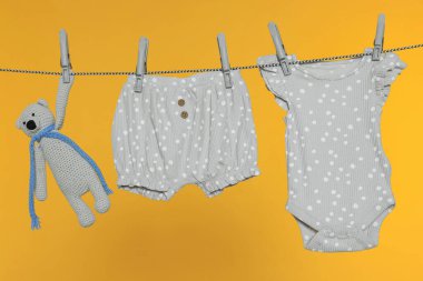 Bebek kıyafetleri ve ayı oyuncağı turuncu arka planda çamaşır ipinde kuruyor.