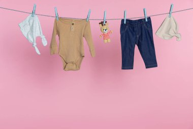 Farklı bebek kıyafetleri ve ayı oyuncağı pembe arka planda çamaşır ipinde kuruyor.