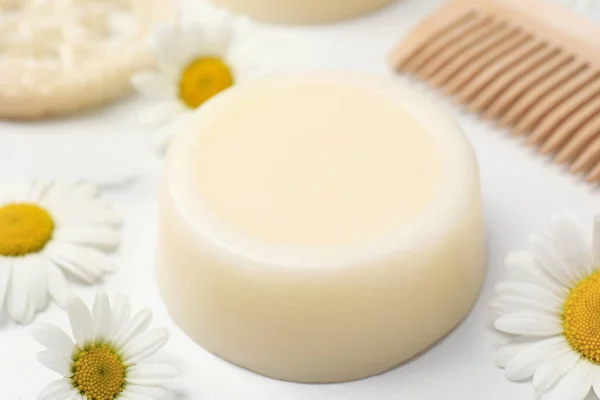 Solid shampoo bar, comb and chamomiles on white table, closeup. Hair care